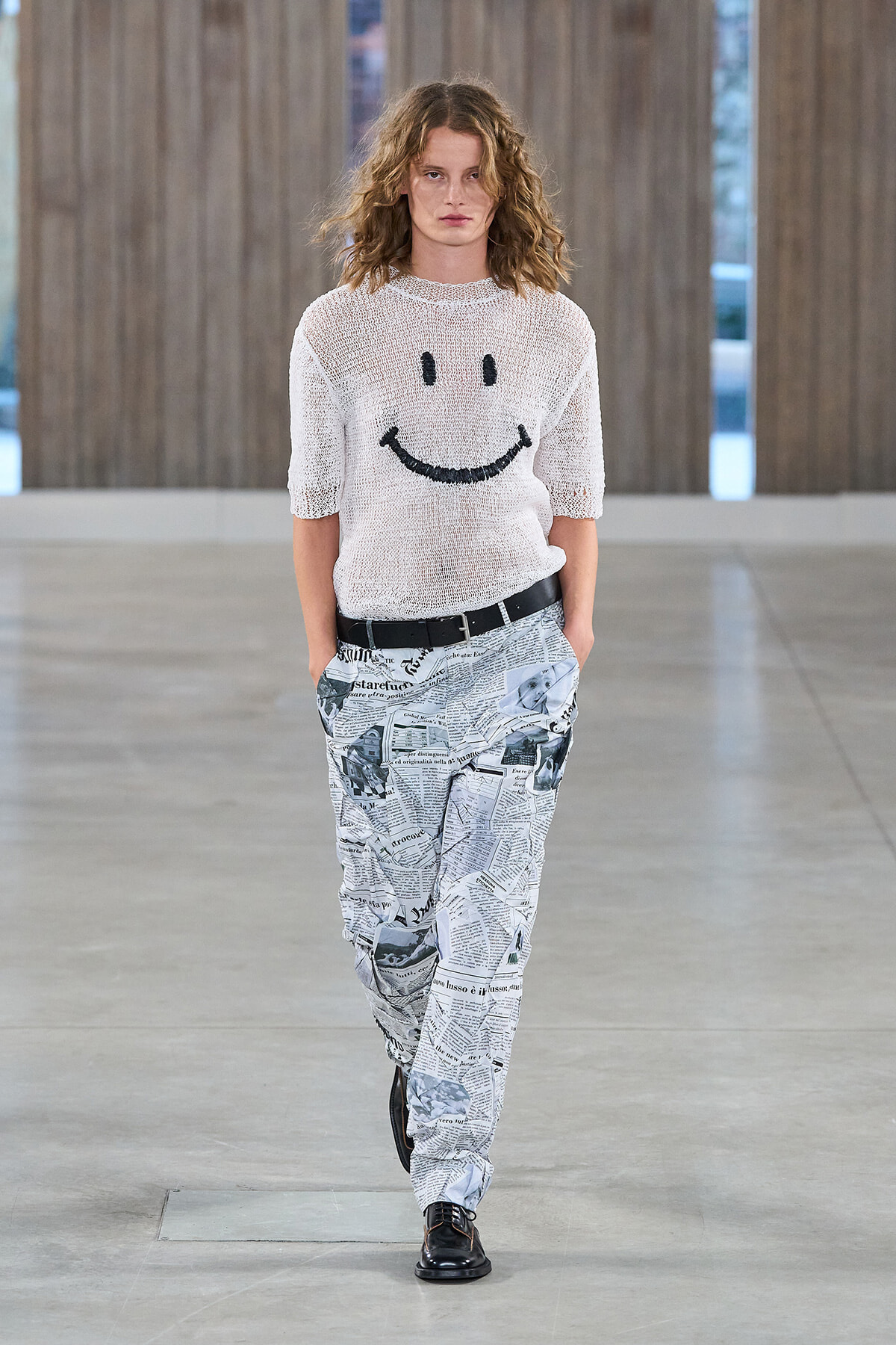 Female fashion model walking a runway in a white knit sweater featuring a black smiley face and newspaper-print pants with a black belt.