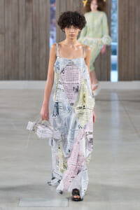 Model walking a runway in a loose, floor-length dress made from newspaper-print fabric, carrying a folded newspaper-inspired bag, indoors on a gray stage.
