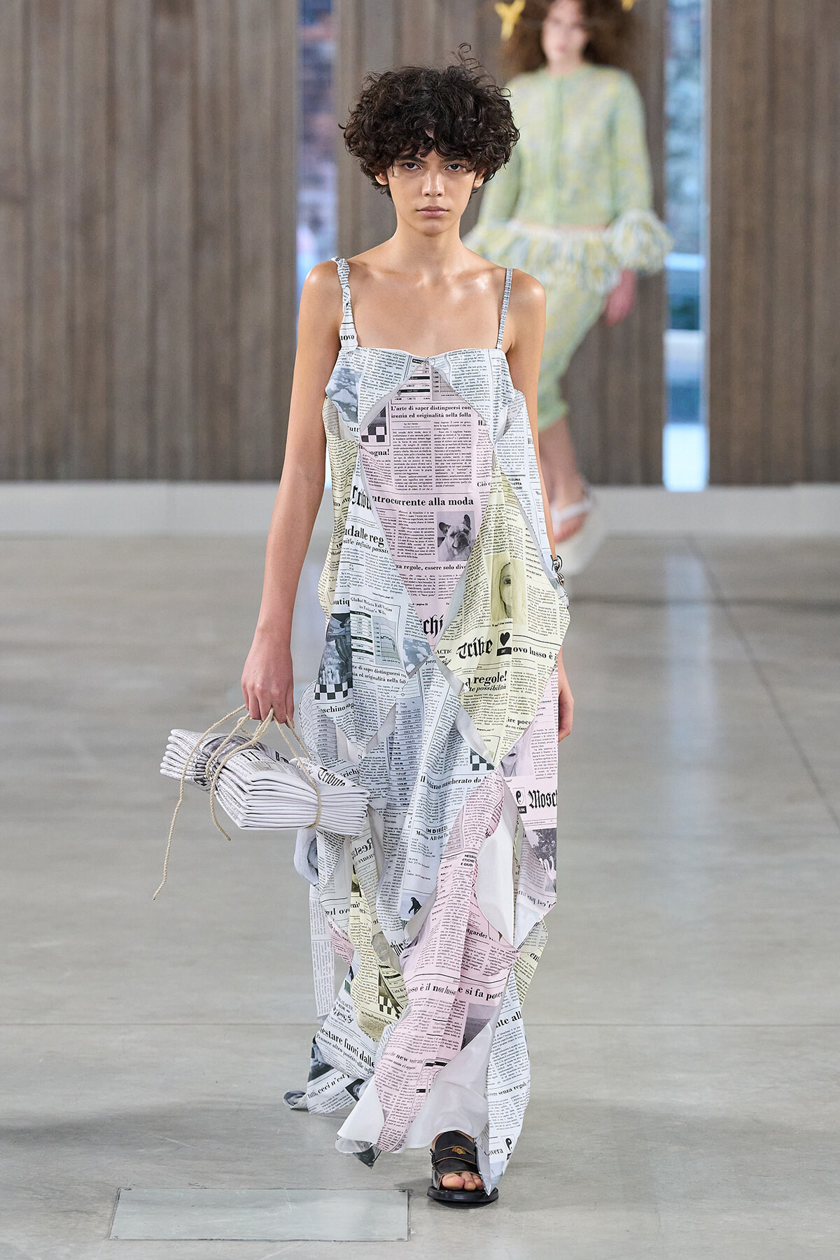 Model walking a runway in a loose, floor-length dress made from newspaper-print fabric, carrying a folded newspaper-inspired bag, indoors on a gray stage.