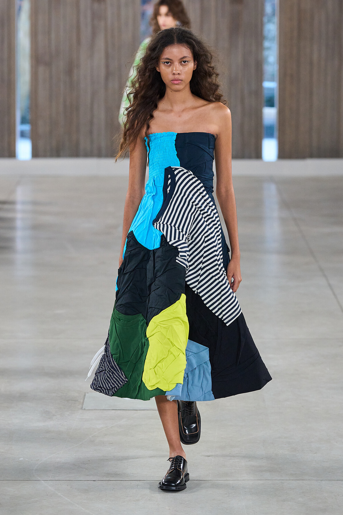 Model walking a fashion runway in a strapless, color-block patchwork gown with blue, green, black, and striped panels, black shoes.