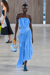 Model walking runway in a bright blue strapless gown with draped bodice, carrying a small blue bucket bag with orange sun design.
