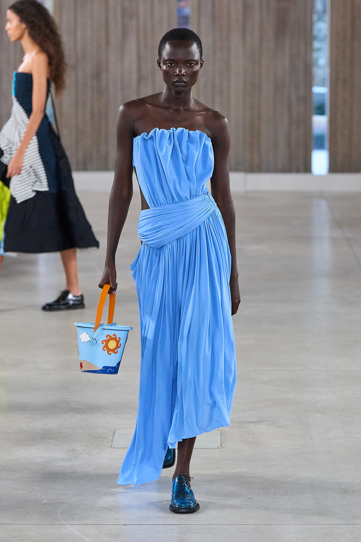 Model walking runway in a bright blue strapless gown with draped bodice, carrying a small blue bucket bag with orange sun design.