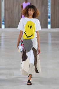 Model walks a runway in a white T-shirt with a large yellow smiley and a patchwork, multicolored skirt with mixed fabrics and black sandals.