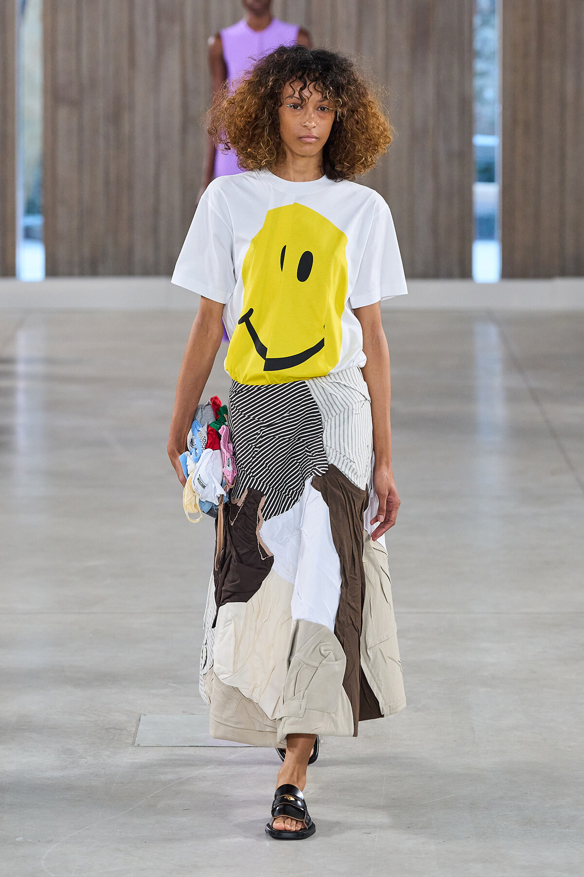 Model walks a runway in a white T-shirt with a large yellow smiley and a patchwork, multicolored skirt with mixed fabrics and black sandals.