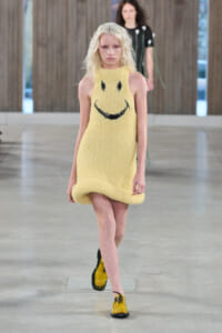 Model walks the runway in a yellow fuzzy sleeveless dress with a large black smiley face, paired with yellow patent shoes and black laces.