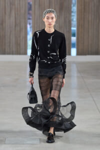Model walks a runway in a black cardigan with white abstract lines, a sheer black striped skirt, black loafers, and a small black bag; white headband and earrings.