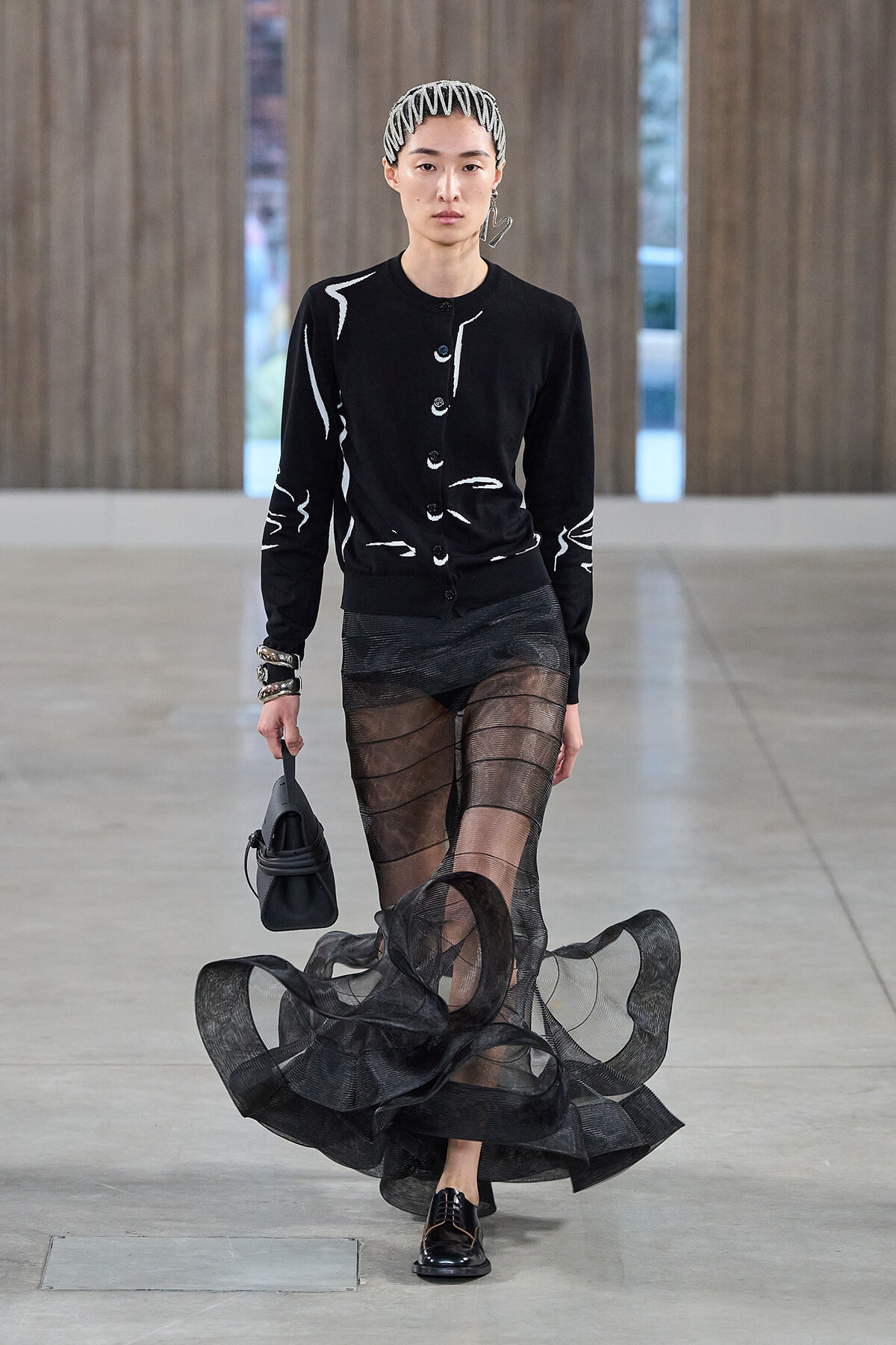 Model walks a runway in a black cardigan with white abstract lines, a sheer black striped skirt, black loafers, and a small black bag; white headband and earrings.