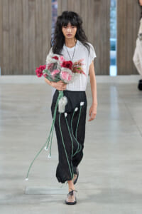 Model walking down a runway holding a large bouquet of pink flowers wrapped in plastic, wearing a white T-shirt and black pants with green stems and white flowers.