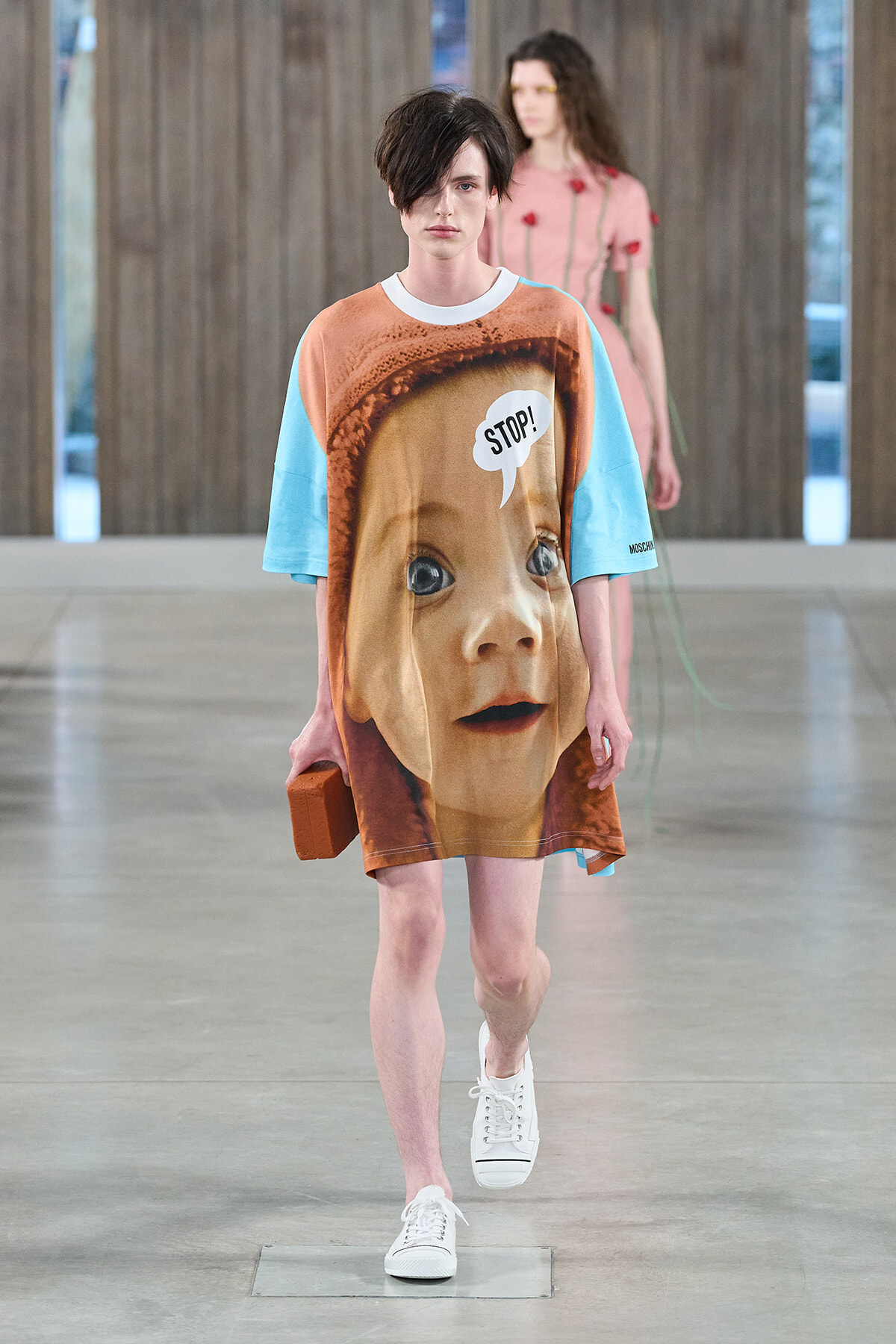 Runway model wearing an oversized dress printed with a giant baby's face and a 'STOP!' speech bubble, blue sleeves, white sneakers, and holding a brick-shaped accessory as another model walks in the background.