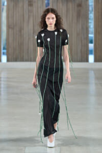 Model walking a runway in a sheer black dress with long green tie cords and white floral appliqués, white heels.