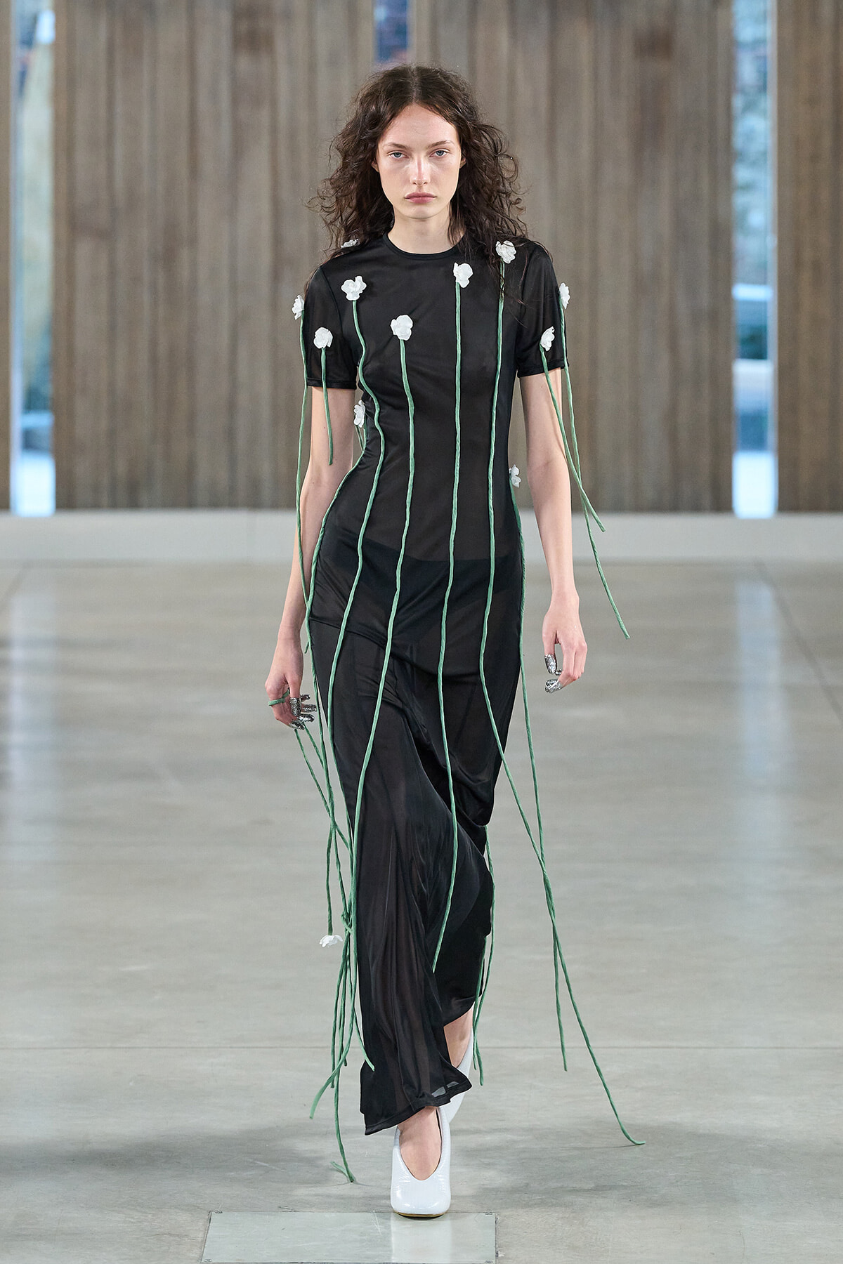 Model walking a runway in a sheer black dress with long green tie cords and white floral appliqués, white heels.