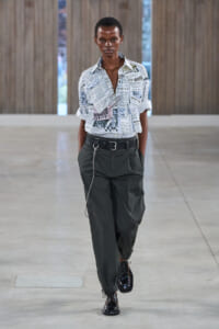 Model walking a runway in a newspaper-print button-down shirt tucked into dark green cargo pants, with a chain belt and black lace-up shoes.