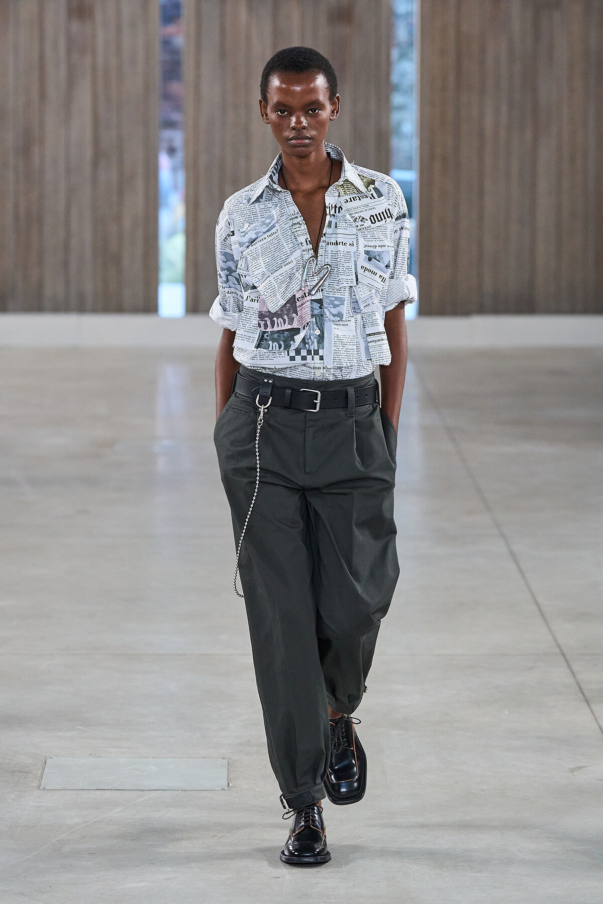 Model walking a runway in a newspaper-print button-down shirt tucked into dark green cargo pants, with a chain belt and black lace-up shoes.