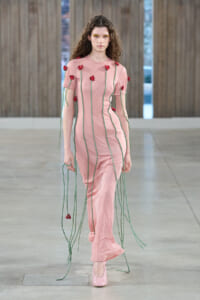 Runway model in a pink dress with vertical green cords and red flower accents, walking on a minimalist catwalk.