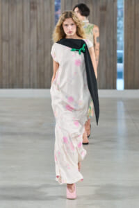 Model walking a fashion runway in a light, pink-splashed maxi dress with a black sash-like scarf and a green balloon-brooch, pink heels visible.