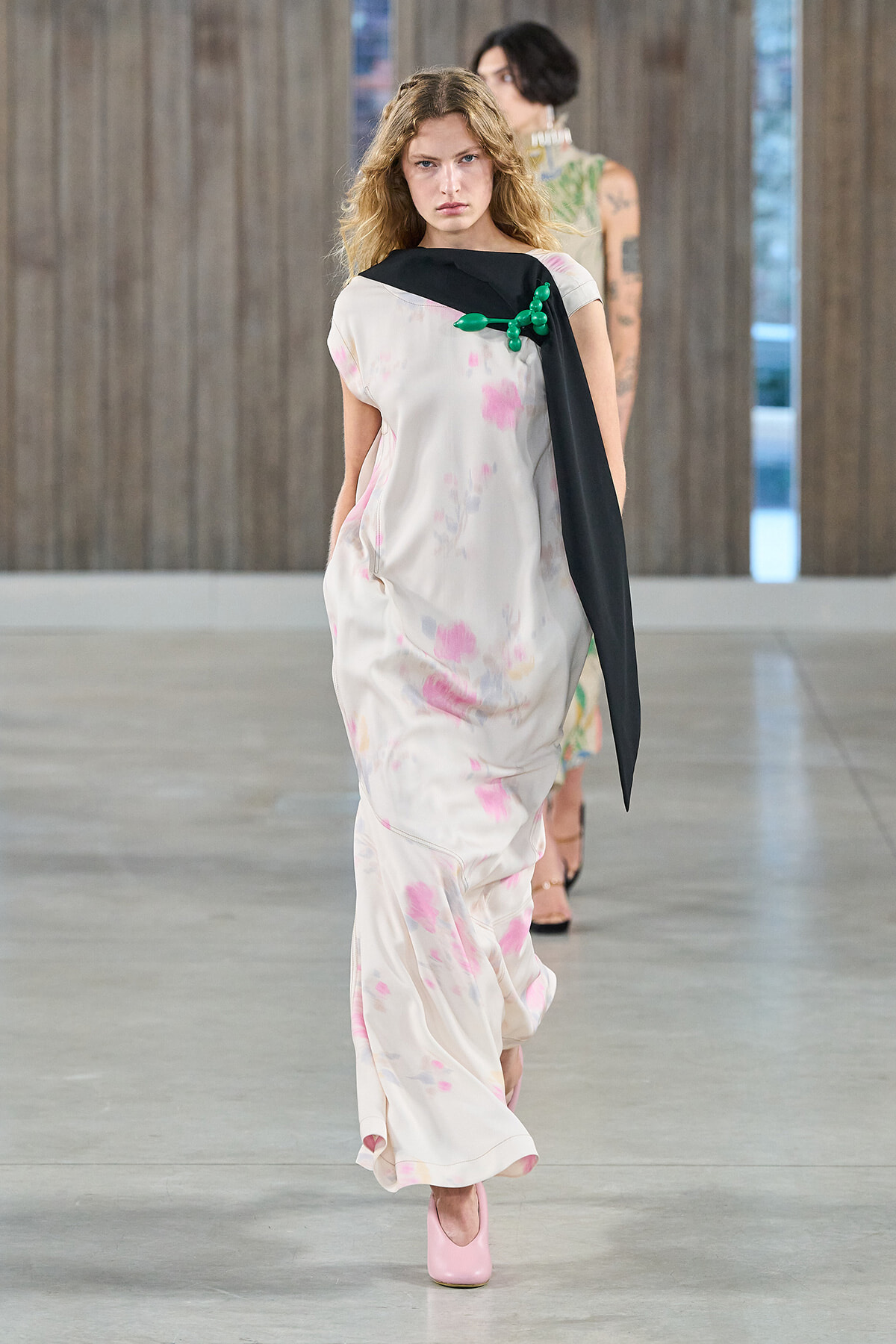 Model walking a fashion runway in a light, pink-splashed maxi dress with a black sash-like scarf and a green balloon-brooch, pink heels visible.