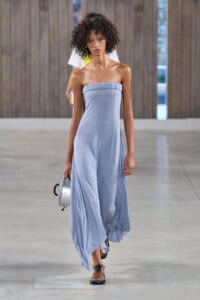 Model walks a fashion runway in a strapless pale blue gown, holding a small silver round handbag with a short handle.