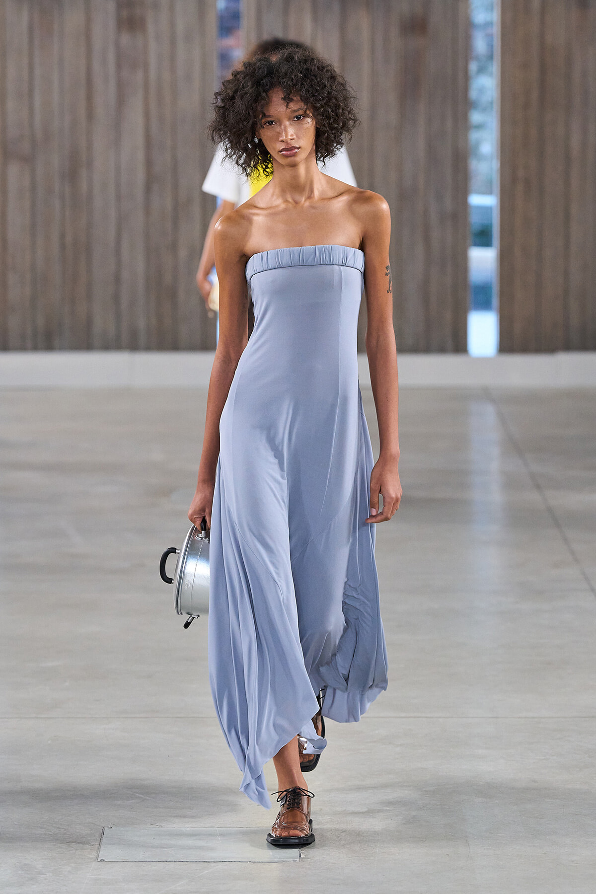 Model walks a fashion runway in a strapless pale blue gown, holding a small silver round handbag with a short handle.