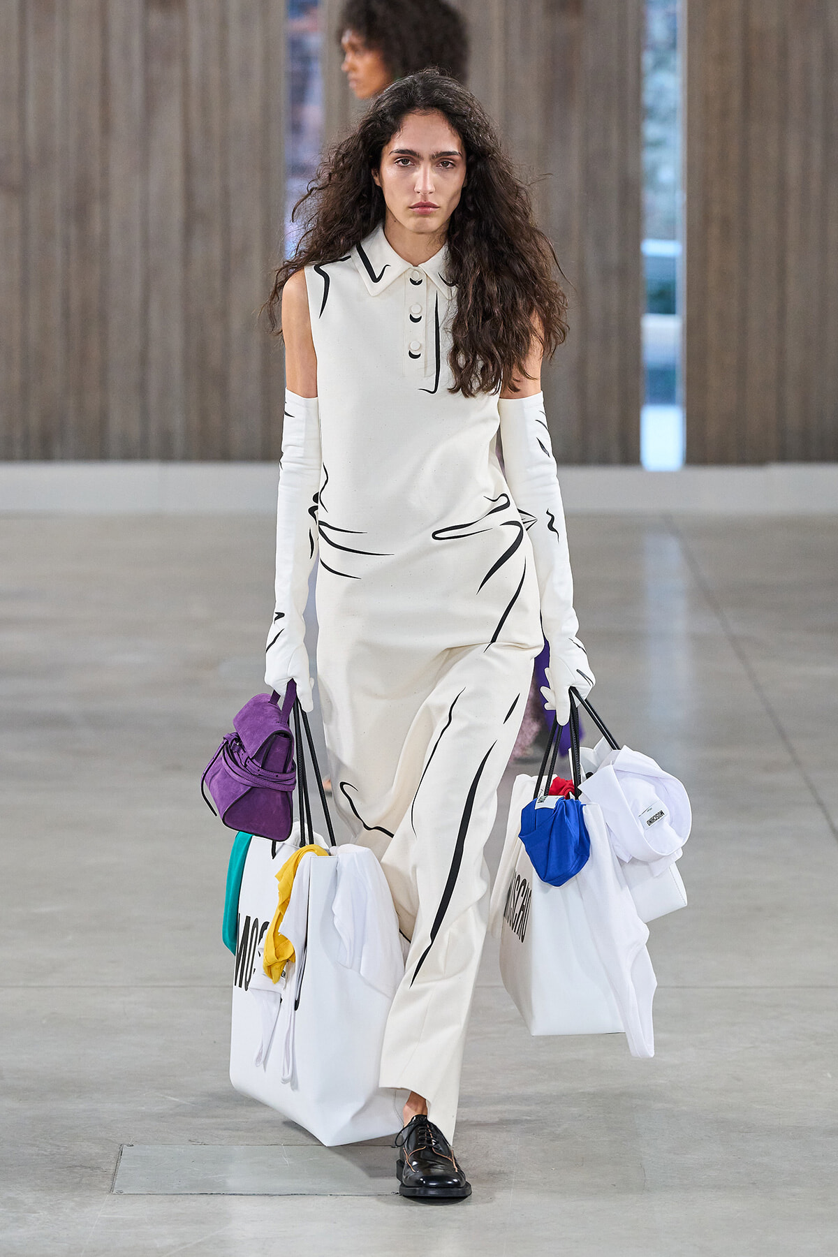 Model walking on a runway in a white sleeveless dress with black abstract lines, carrying several large white shopping bags and colorful accessories.