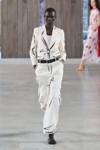 Model wearing a white blazer and wide-leg pants with black abstract stripes, belt at the waist, walking on a runway.