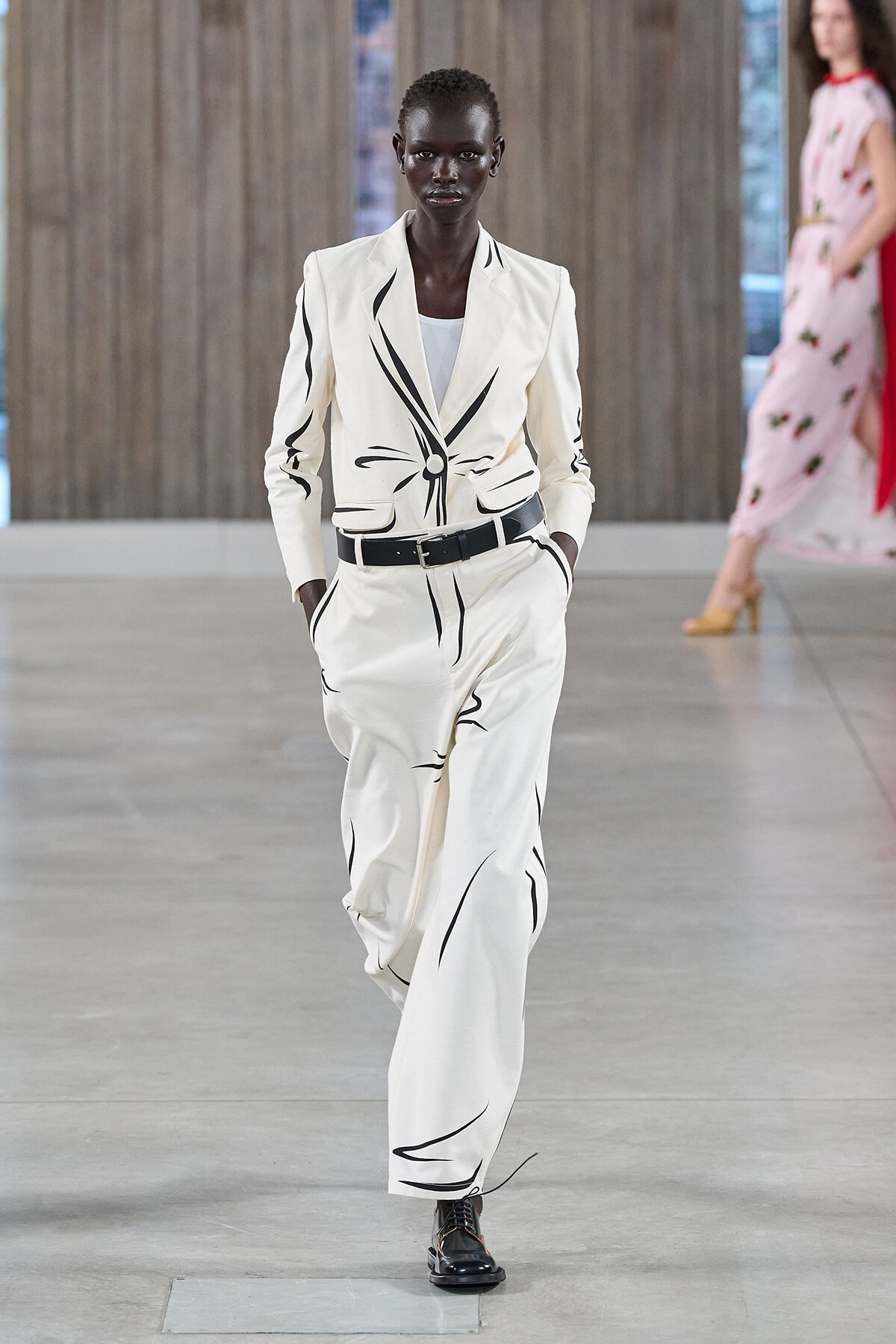 Model wearing a white blazer and wide-leg pants with black abstract stripes, belt at the waist, walking on a runway.