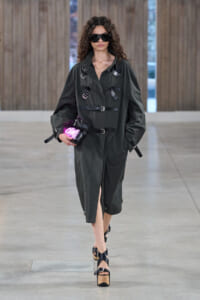 Model in a dark green belted trench with multiple buckles, wearing large black sunglasses and carrying a pink neon clutch on a runway.