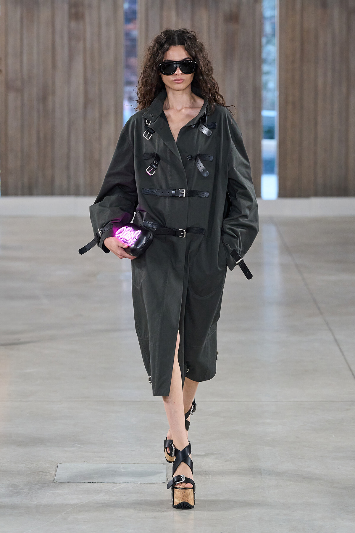 Model in a dark green belted trench with multiple buckles, wearing large black sunglasses and carrying a pink neon clutch on a runway.