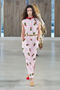 Female model walking a fashion runway in a pale pink sleeveless jumpsuit with red collar and scattered green cherry prints, cinched with a beige belt.