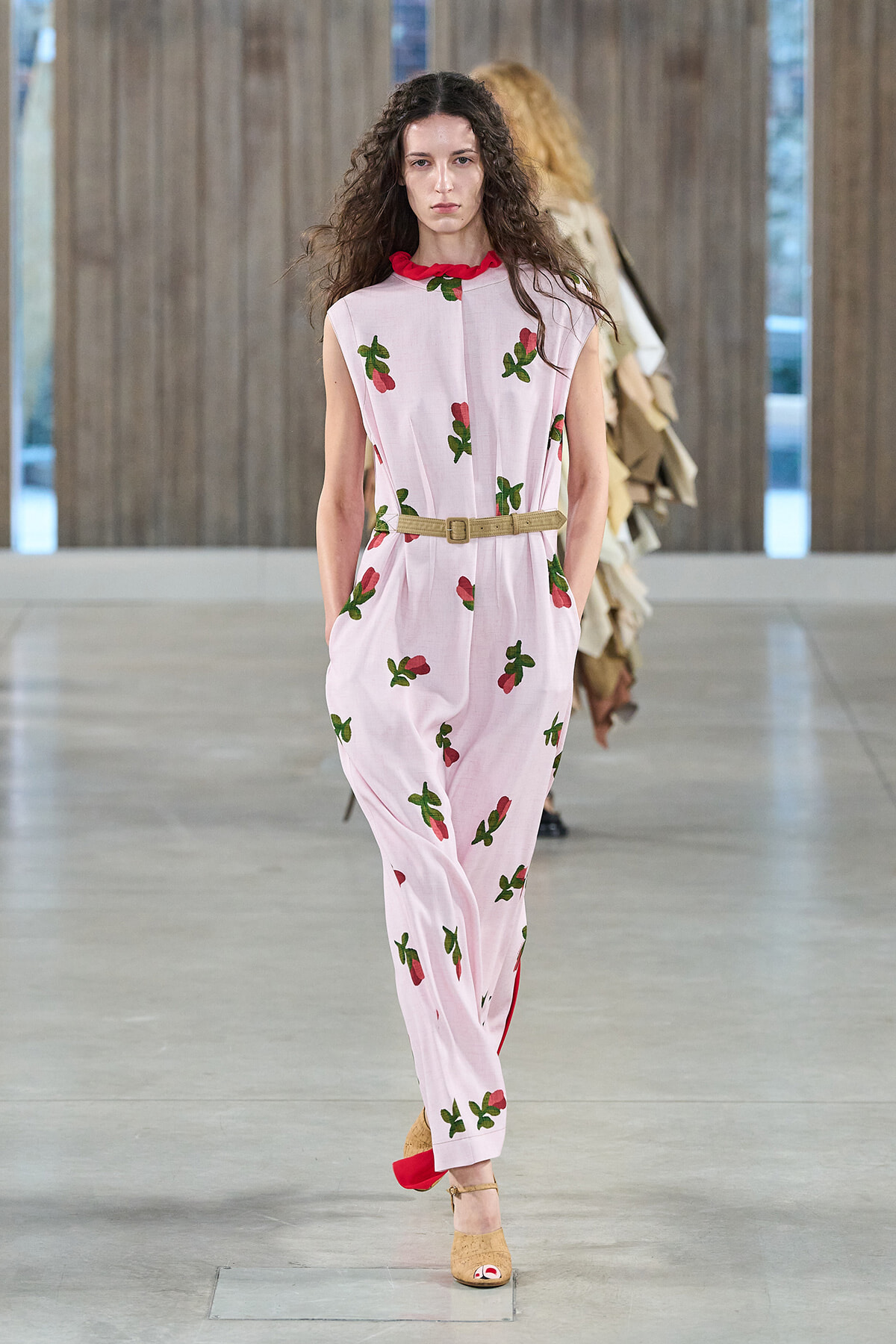Female model walking a fashion runway in a pale pink sleeveless jumpsuit with red collar and scattered green cherry prints, cinched with a beige belt.
