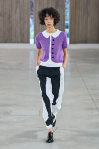 Model walking a runway in a purple knit cardigan with a white collar and black-and-white color-block pants, black loafers visible.