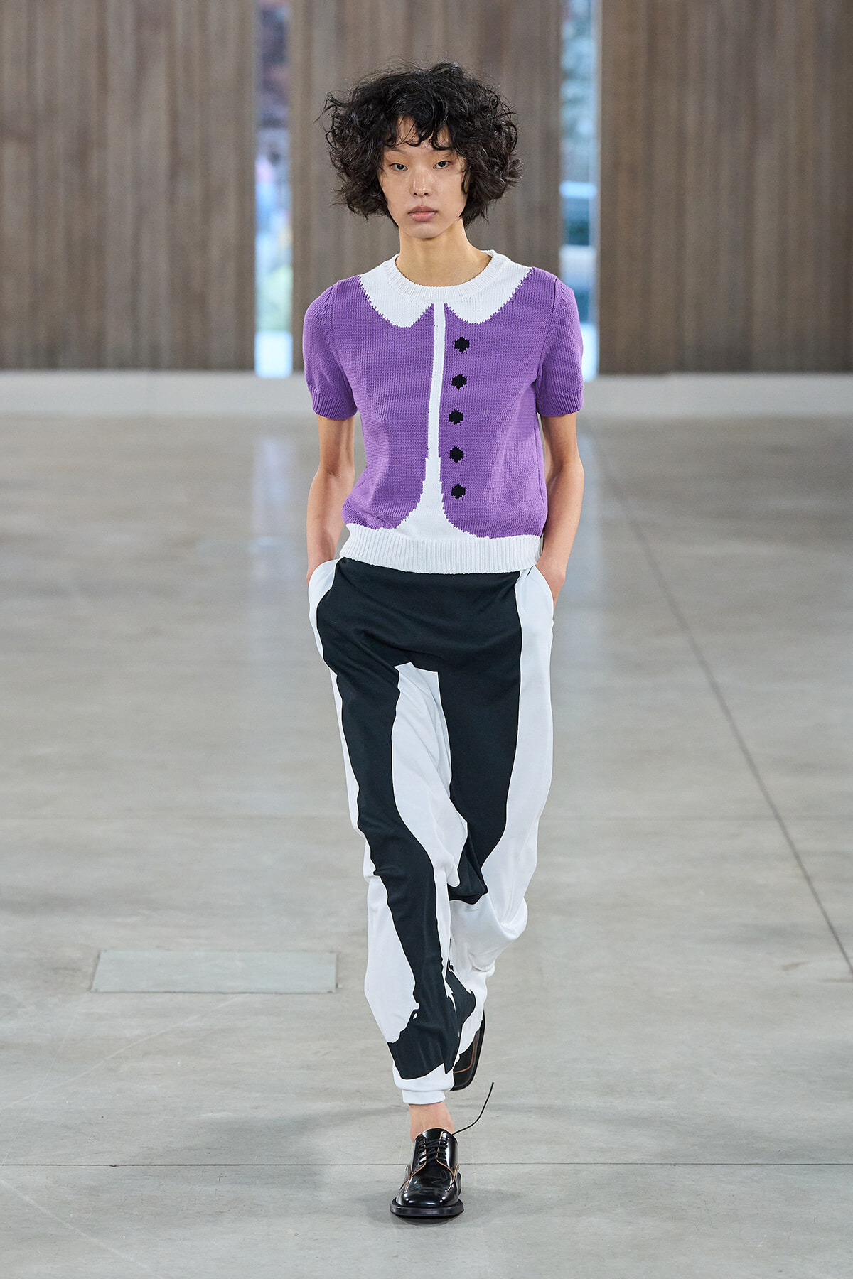 Model walking a runway in a purple knit cardigan with a white collar and black-and-white color-block pants, black loafers visible.