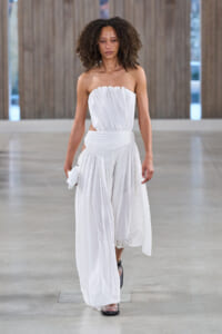 Model walks a runway in a white strapless, draped gown with a wrapped waist and flowing skirt, holding a small white clutch in one hand.