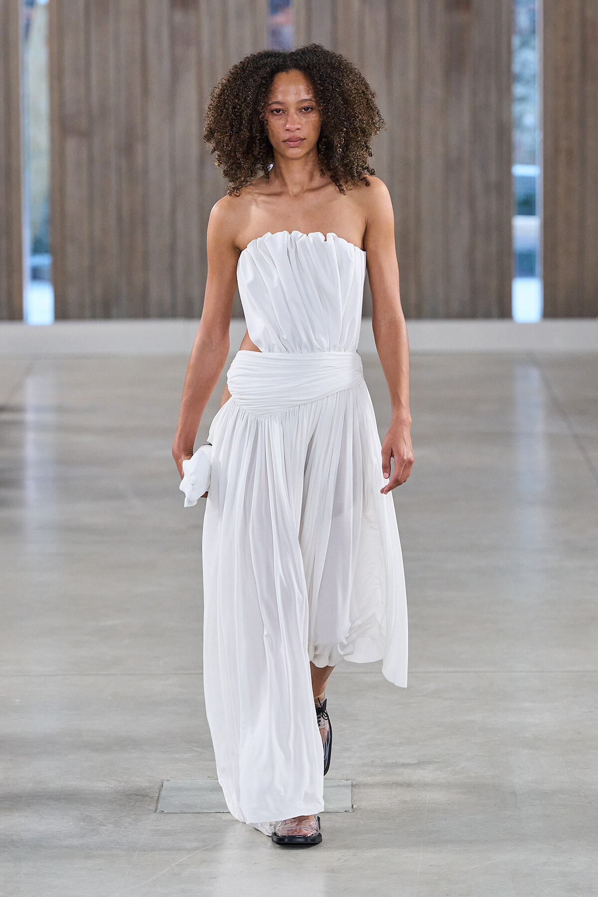 Model walks a runway in a white strapless, draped gown with a wrapped waist and flowing skirt, holding a small white clutch in one hand.