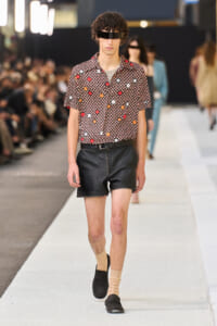 Male model walks the runway in a short-sleeve floral print shirt and black leather shorts, beige socks, and black loafers, with a black visor accessory.
