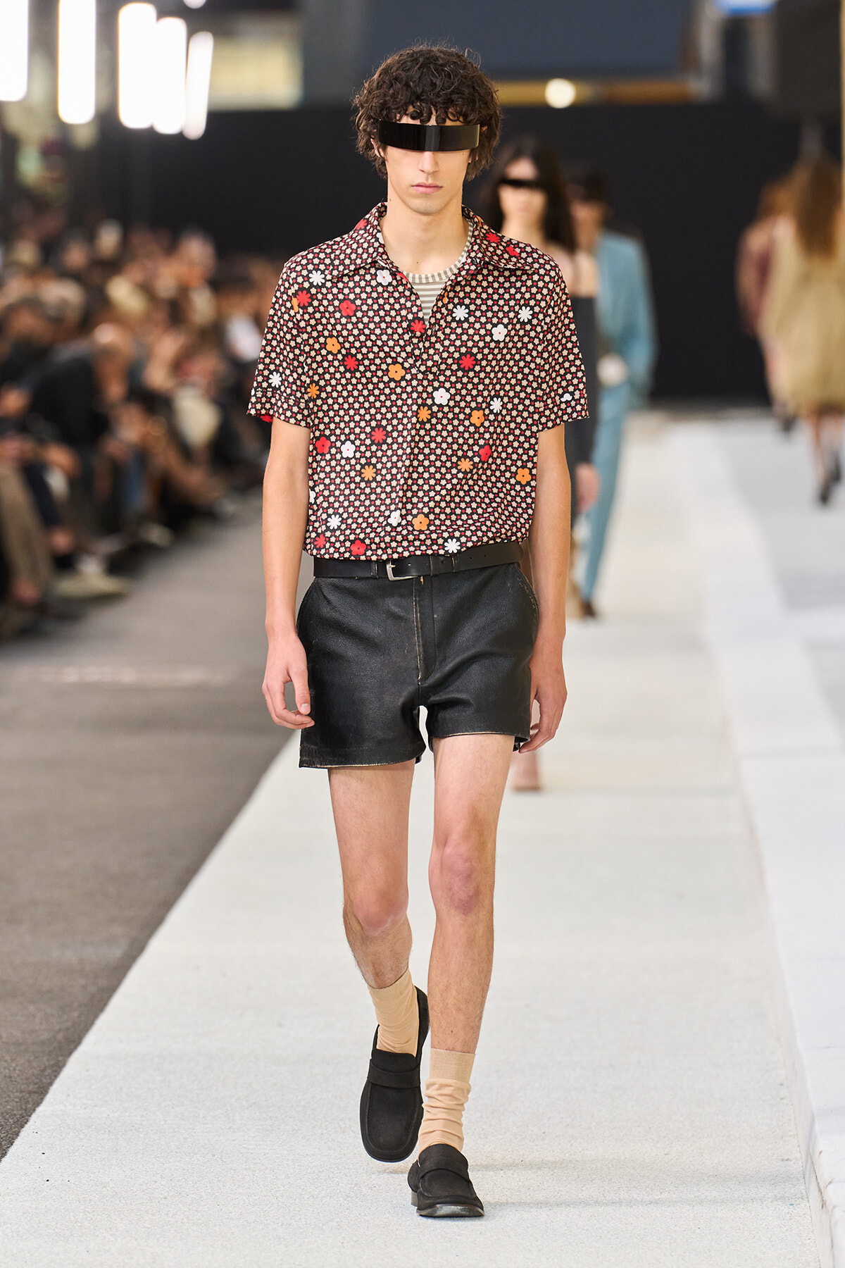 Male model walks the runway in a short-sleeve floral print shirt and black leather shorts, beige socks, and black loafers, with a black visor accessory.