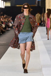 Model walks the runway wearing a long floral-patterned coat over an olive green knit sweater and light blue shorts, with tan socks and brown loafers; eyes obscured by a black blindfold.