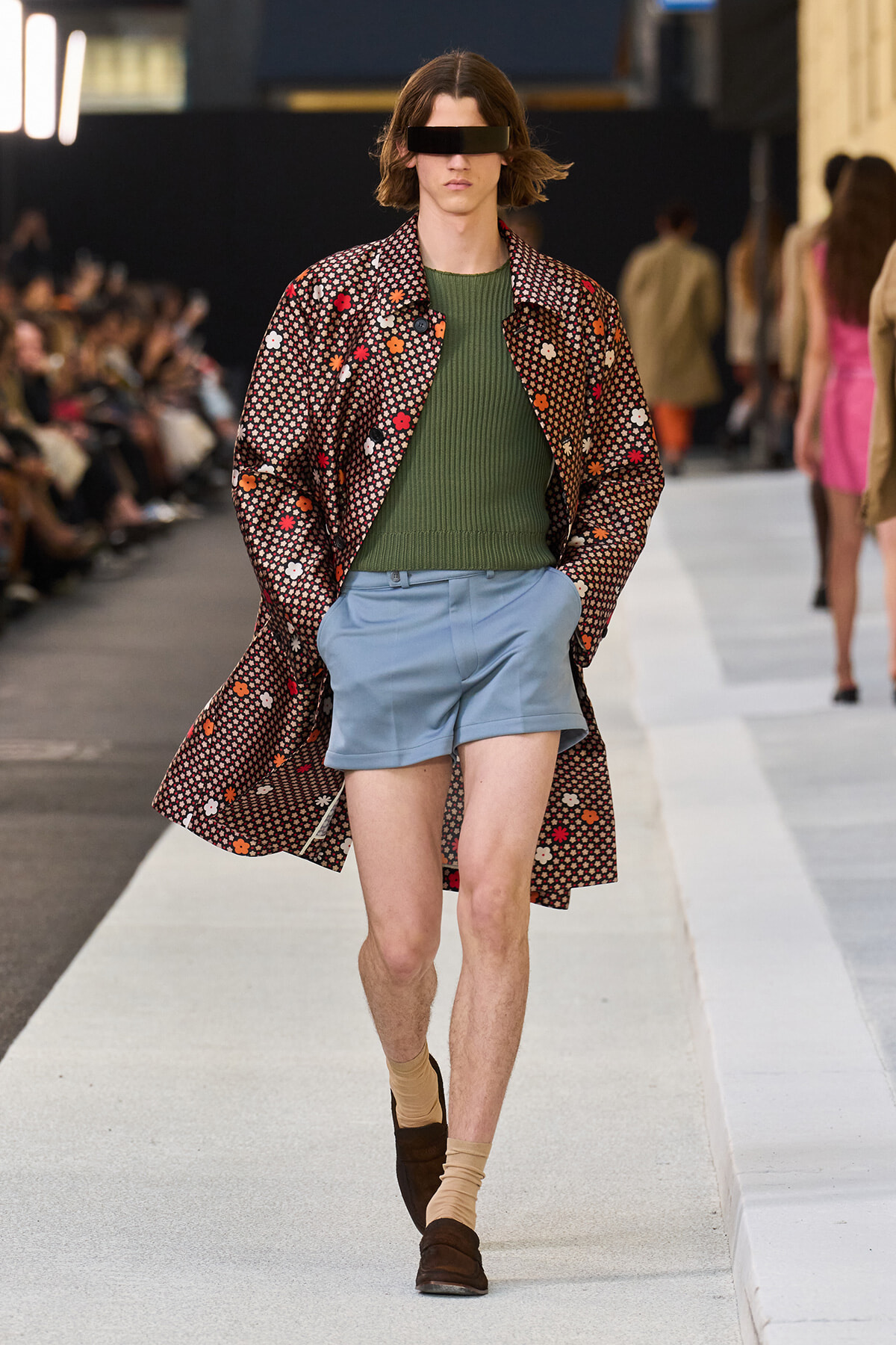 Model walks the runway wearing a long floral-patterned coat over an olive green knit sweater and light blue shorts, with tan socks and brown loafers; eyes obscured by a black blindfold.
