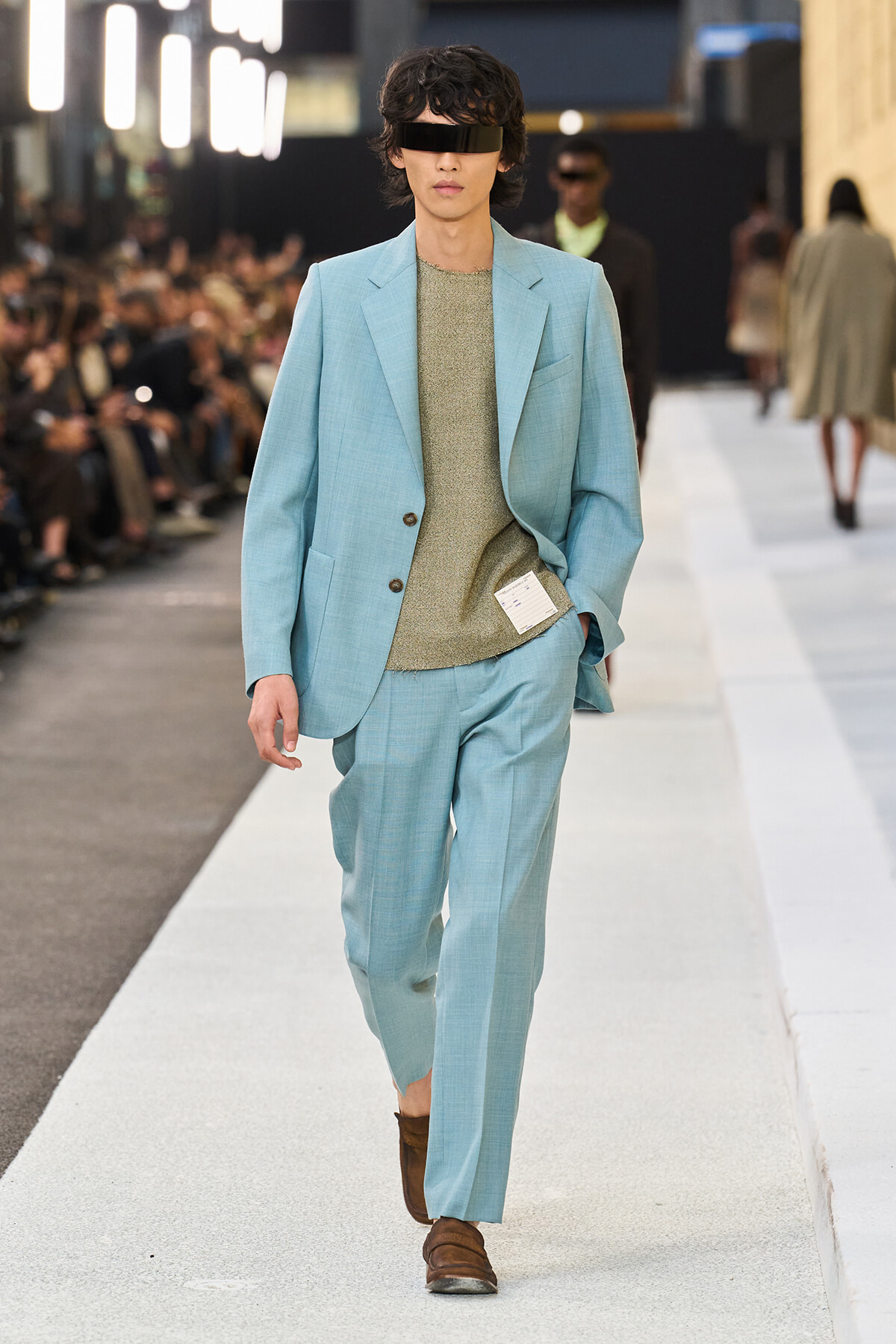 Male model walks a fashion runway in a pale blue suit with a beige knit top and brown shoes, wearing a black visor-style eye mask.