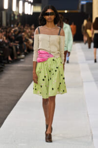 Runway model wearing a beige off-shoulder top with sheer neckline, pink belt, and a green floral midi skirt with brown loafers and oversized sunglasses.
