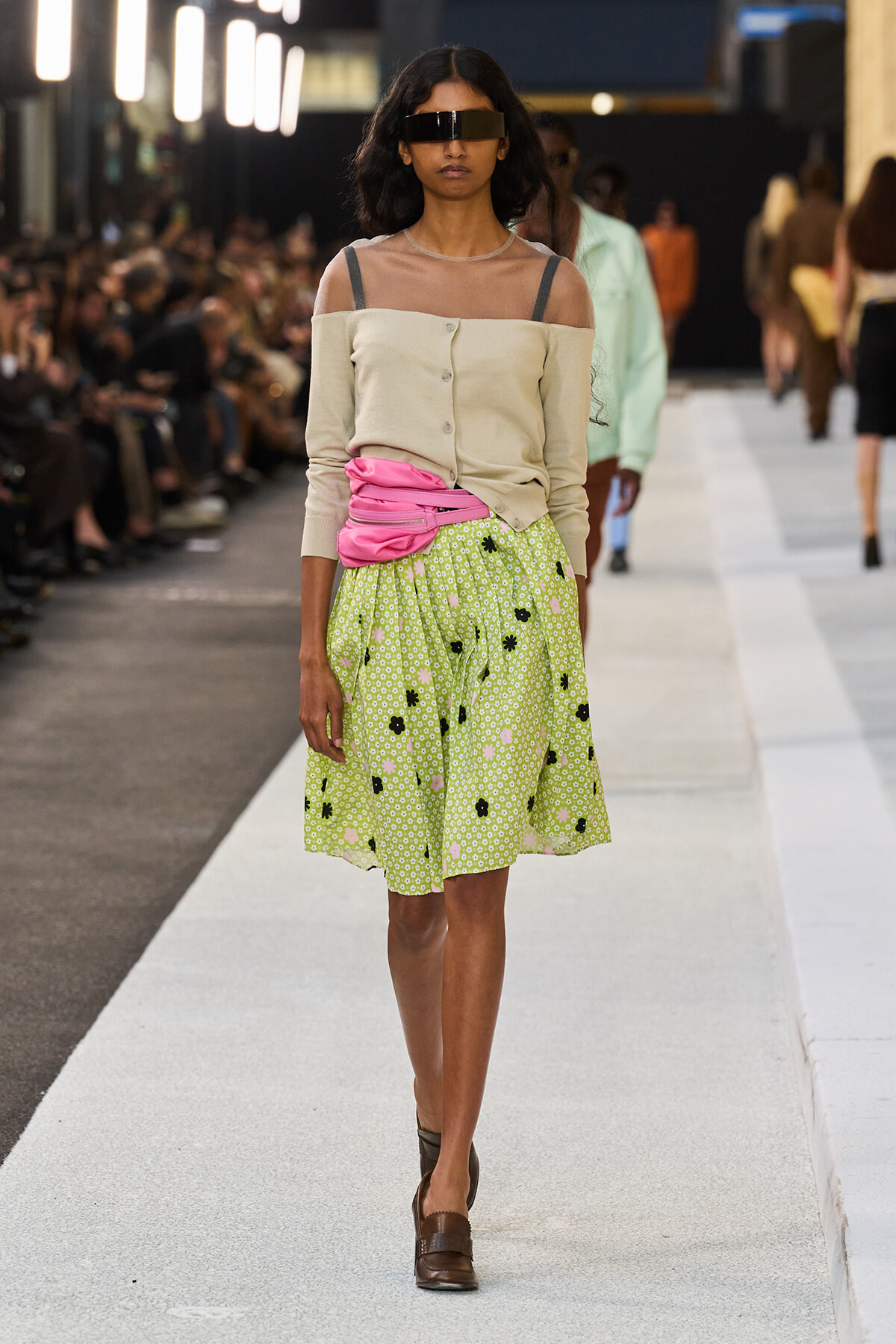 Runway model wearing a beige off-shoulder top with sheer neckline, pink belt, and a green floral midi skirt with brown loafers and oversized sunglasses.