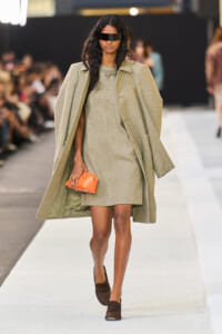 Model on a runway wearing a beige textured coat over a matching mini dress, carrying an orange clutch and wearing a black visor sunglasses.
