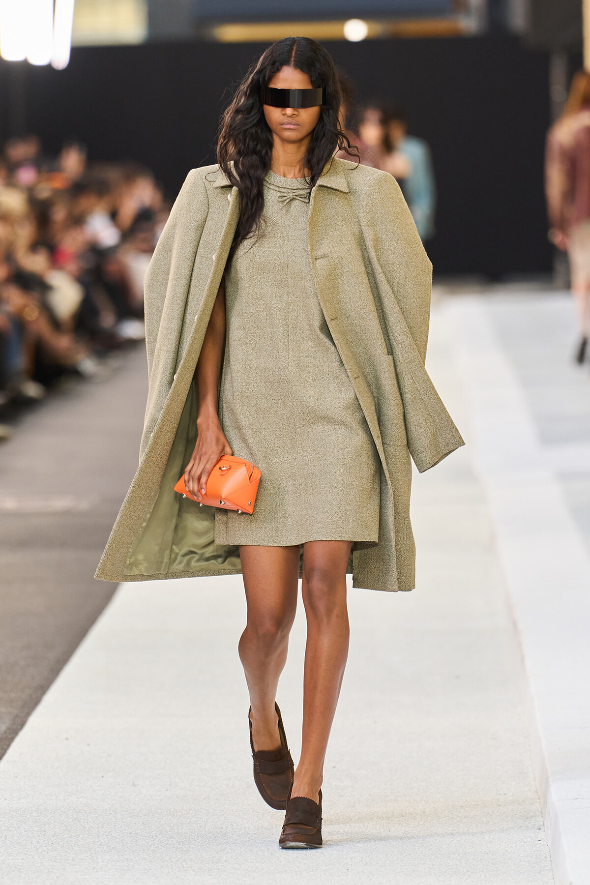 Model on a runway wearing a beige textured coat over a matching mini dress, carrying an orange clutch and wearing a black visor sunglasses.