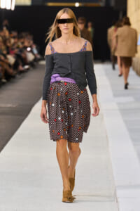 Model walking a fashion show wearing a dark off-shoulder cardigan over a floral-strap top, a purple waist sash, and a black floral skirt, with tan loafers and a futuristic visor sunglasses piece.