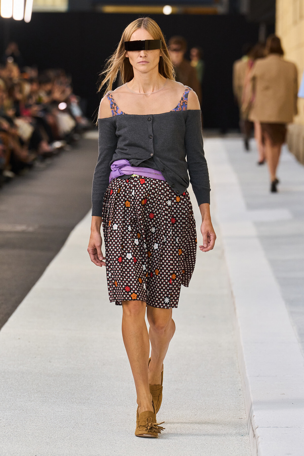 Model walking a fashion show wearing a dark off-shoulder cardigan over a floral-strap top, a purple waist sash, and a black floral skirt, with tan loafers and a futuristic visor sunglasses piece.