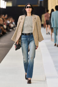 Model on a runway wearing a beige blazer over a gray knit top, distressed jeans, a black belt, and dark ankle boots with futuristic visor sunglasses.