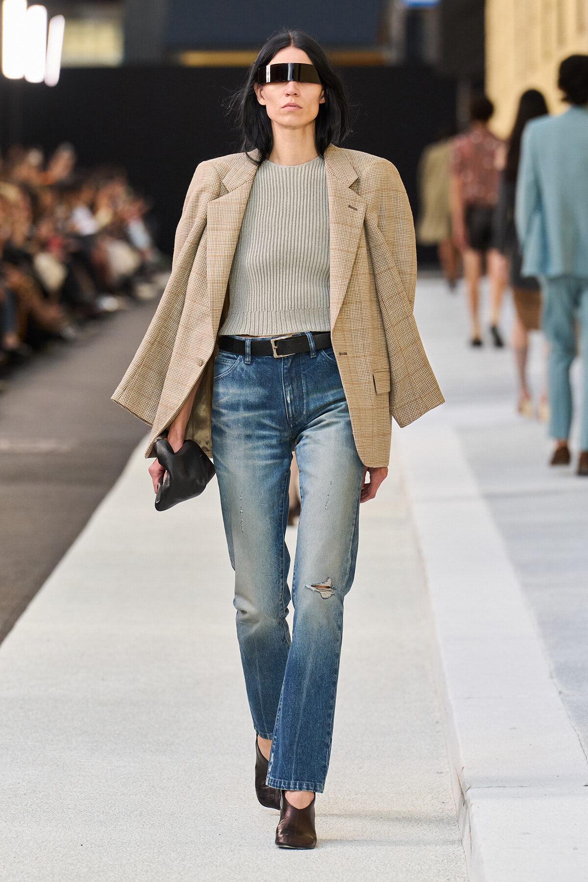 Model on a runway wearing a beige blazer over a gray knit top, distressed jeans, a black belt, and dark ankle boots with futuristic visor sunglasses.