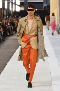 Runway model wearing a tan trench over a beige suit and bright orange pants with an orange sash belt, eyes covered by a black blindfold.