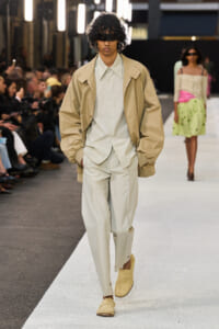 Male model walking a fashion runway in a beige oversized jacket over a light suit with beige loafers and a black eye mask/blindfold.