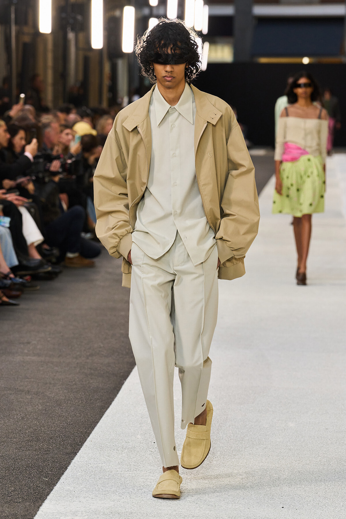 Male model walking a fashion runway in a beige oversized jacket over a light suit with beige loafers and a black eye mask/blindfold.