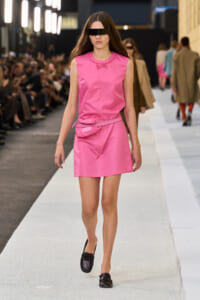Model walking the runway in a bright pink sleeveless dress with a belt bag and black visor sunglasses.